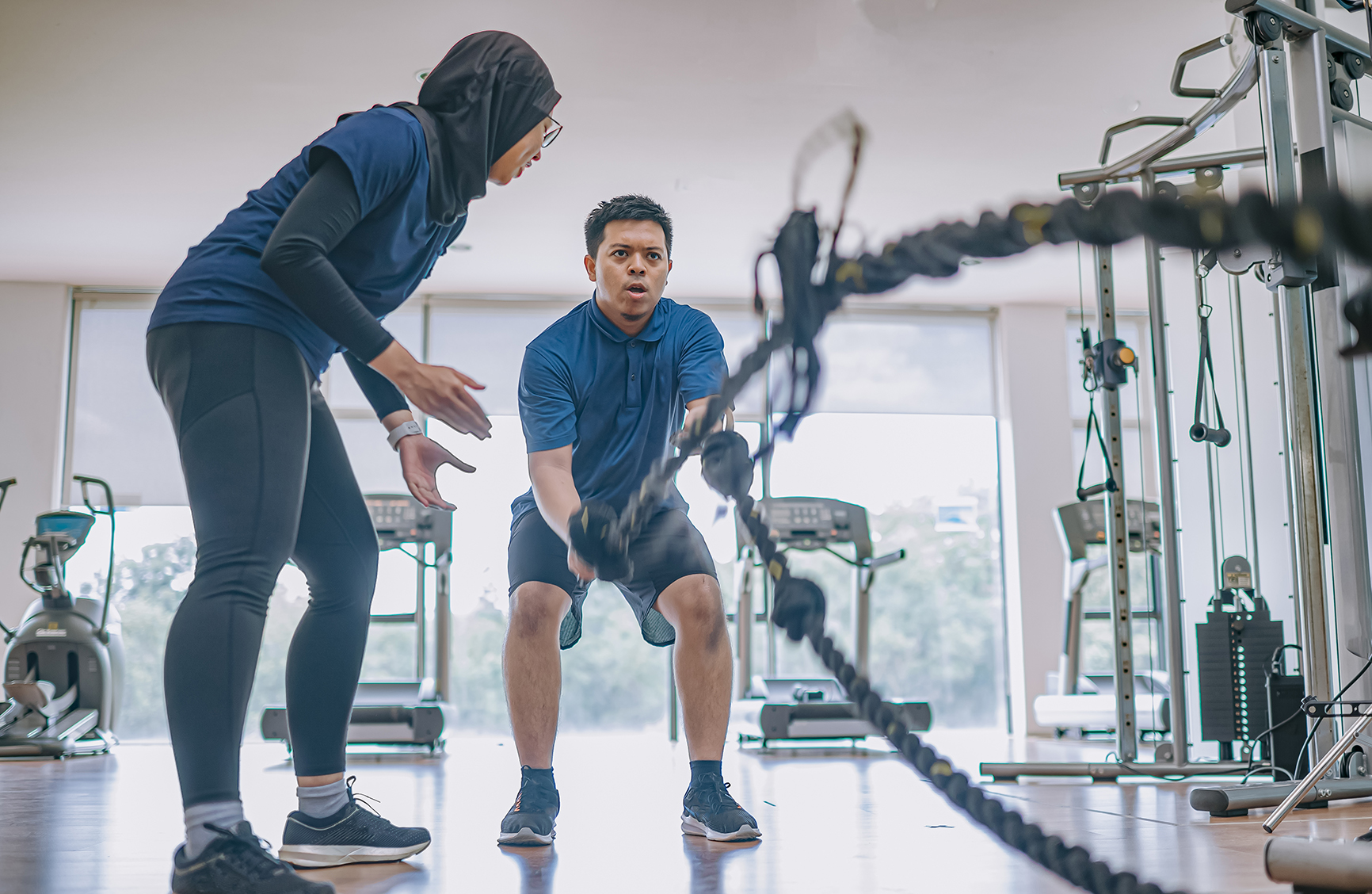 Asian Malay female personal trainer encouraging Asian Malay man practicing challenging battle rope in gym